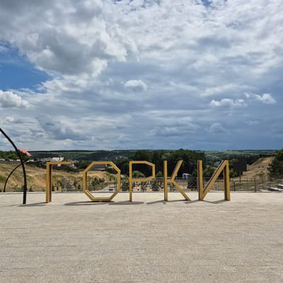 In the city of Liski, Russia, an outdoor scene features large, golden, block-style letters spelling out "ГОРКИ" (GORKI) in the foreground. The letters are positioned on a light-colored, textured stone pavement. Behind the letters, a landscaped area with a retaining wall and some vegetation is visible, leading up to a hillside. Further in the background, a small town or village with houses and trees can be seen nestled in a valley. The sky above is filled with dramatic, grey and white clouds against a blue sky. The overall impression is of an urban park or recreational area.