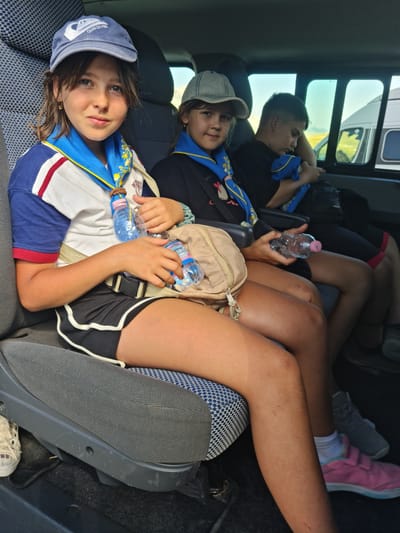 Three young people are seated in the back of a van. The person in the foreground on the left is a female wearing a blue cap, a scout scarf, a white shirt with blue and red sleeves, and black shorts. She is holding two plastic water bottles. Beside her, to the right, is another female wearing a grey cap and a scout scarf. She is holding a plastic water bottle. Behind them, a male is seated, wearing a black t-shirt and black shorts. He is looking down at his phone and holding a blue object. The interior of the van has grey seats with a blue and white patterned fabric on the side. Outside the windows, a landscape with hills is visible. The image does not provide specific details to ascertain the location within the city of L'Aquila, Italy, beyond being in the rear of a vehicle with outdoor scenery.