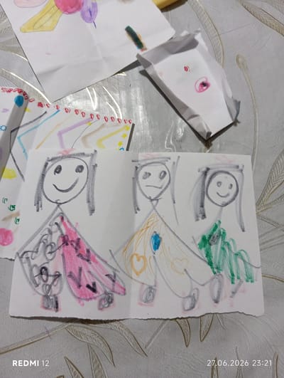 The image shows a close-up, high-angle view of several children's drawings on a textured, light-colored surface, possibly a table. The drawings are scattered and layered, indicating they are on display. One drawing in the foreground depicts three stick-figure people with simplified facial features and colorful dresses. The person on the left wears a pink dress with dark markings, the middle person a yellow dress with a blue jewel, and the person on the right a green dress. Another drawing above shows abstract shapes and colors, including yellow, pink, and purple, possibly representing a sun or abstract figures. A folded piece of paper with smudged pink and red markings is visible in the upper right. The overall scene is a candid snapshot of children's artwork. The bottom of the image displays the text "REDMI 12" on the left and "27.06.2026 23:21" on the right, indicating the device and date/time of capture. The environment does not provide any specific visual cues to determine its location in Bekabad, Uzbekistan.