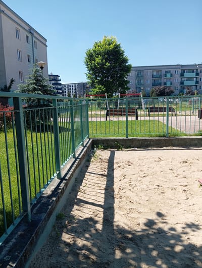 The image shows a residential area in Radzymin, Poland, on a clear, sunny day. In the foreground, a sandpit is visible, bordered by a low concrete wall and a green metal fence. Beyond the fence, a well-maintained lawn stretches out, with several park benches and playground equipment, including a swing set, suggesting a communal recreational space. In the background, modern apartment buildings are visible, some with balconies and light-colored facades. A large, leafy tree stands prominently in the middle ground, casting shadows on the lawn and the sandpit. The overall scene depicts a peaceful, urban environment with amenities for residents.