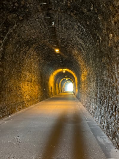 The image shows the interior of a tunnel in Solingen, Germany. The tunnel is constructed of rough-hewn stone, with a smooth, possibly concrete or metal, structure on the right side of the image. The tunnel curves towards a bright exit in the distance. Dim, yellow lights are spaced along the left wall. A metal safety railing is also visible on the left wall. The floor appears to be cobblestone or similarly paved stone. No people are visible.