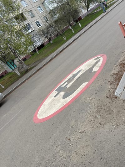 The image shows a street in Sayansk, Russia. The street has a faded road marking depicting pedestrians crossing. A multi-story residential building is visible on the left, partially obscured by trees. Two people are seen walking along the side of the road. The street surface is gray asphalt with some dirt and debris.
