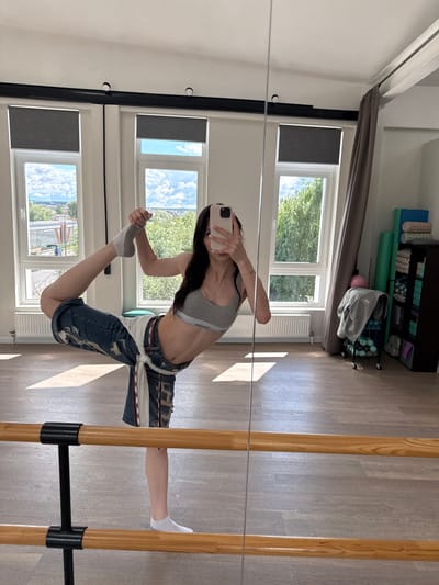 A young woman is captured in a full-body mirror selfie, performing a split with her right leg extended forward and her left leg bent behind her. She is wearing a grey sports bra with "Calvin Klein" visible on the band, distressed denim shorts tied with a white belt, and grey and white socks. Her right hand is extended upwards, holding her phone to take the photo, while her left hand is bent and reaching towards her left foot. The setting appears to be a dance or fitness studio, indicated by the wooden floor, barre, and large windows. Sunlight streams in from the windows, casting shadows on the floor. Through the windows, trees and a bright blue sky with scattered clouds are visible, suggesting daytime and fair weather. In the background, shelves are stocked with various fitness equipment, including yoga mats and foam rollers. The overall mood is one of athleticism and flexibility. The image is oriented vertically, suggesting it was taken in portrait mode.