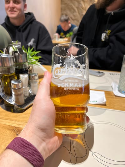 A close-up image taken in a restaurant or bar in Niš, Serbia, shows a right hand, wearing a maroon braided wristband, holding a clear glass. The glass is branded "Carlsberg, COPENHAGEN DENMARK, 1847 ONWARDS" and contains a golden amber liquid, consistent with beer, with residual foam and condensation adhering to the upper interior surface. The liquid level is approximately one-third full. The hand is positioned over a light-colored placemat with a minimalist black line design, resting on a light-colored wooden table.
To the left of the hand, on the table, is a silver-colored condiment caddy containing four clear glass bottles with silver caps. These appear to hold olive oil, balsamic vinegar, salt, and pepper. A small stack of white paper napkins is also visible in the caddy.
In the mid-ground, seated at the table, are two men. The man on the left, partially in frame and smiling, wears a dark hooded sweatshirt. The man on the right, mostly obscured by the beer glass, also wears a dark hooded sweatshirt, with a portion of his beard visible. Further in the blurry background, a third individual with short, curly hair is visible, leaning over a table, potentially eating from a bowl. The background wall features a textured, light-colored stone or stucco finish. A silver smartphone is visible on the table to the right of the beer glass.