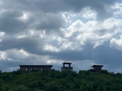 The image depicts a series of modern, angular buildings situated atop a densely forested hill. The sky above is filled with dramatic, dark, and grey clouds, suggesting overcast or approaching stormy weather. The buildings appear to be constructed with a combination of stone or brick and dark-colored framing. The largest structure on the left is long and horizontal with visible windows. To its right, a taller, more open structure with a distinct roofline stands, followed by another building with a similar roof style on the far right. The foliage on the hill is a deep green and appears lush. The image is taken from a distance, and the overall scene is somewhat hazy, potentially due to atmospheric conditions or the image quality. The location of Щебетовский поселковый совет, Ukraine is not visibly indicated in the image itself.