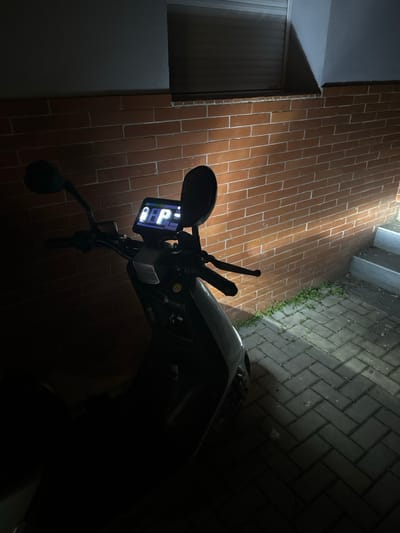 The scene depicts a dark-colored scooter parked at night in Holtzheim, France. The scooter is positioned on gray interlocking pavers, laid in a herringbone pattern, with a reddish-brown brick wall directly behind it. The top-left portion of the image shows a white-framed window with a closed roller shutter above the brick wall. A strong artificial light source, possibly emanating from the scooter itself or an external fixture, illuminates the brick wall, the pavers, and a set of concrete steps rising on the right side of the frame. A small patch of green vegetation is visible at the base of the wall near the steps. The scooter's digital dashboard is illuminated, displaying a fuel gauge icon and a prominent capital letter "P," indicating a parking or park mode status. A yellow circular sticker is affixed to the scooter's frame below the handlebars. The lower and left portions of the image are enveloped in deep shadow. No individuals or active interactions are observed.