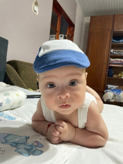 A baby is lying on its stomach on a white surface, wearing a white tank top and a blue and white striped hat. The baby has light skin and blue eyes, and is looking up at the camera with a neutral expression. The background is blurred but appears to be a room with wooden furniture. A red string is tied around the baby's left wrist. The scene is brightly lit, suggesting daytime. The overall impression is one of a relaxed and intimate moment.