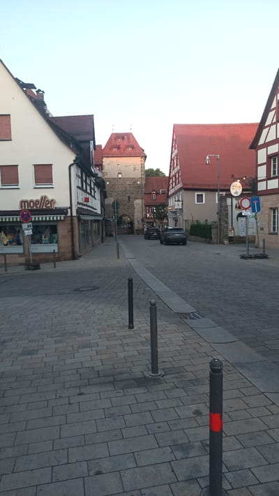 This is a medium shot, taken from street level, of a picturesque street in Altdorf bei Nürnberg, Germany. The scene captures the charming architecture of the old town, with a prominent gatehouse or tower at the end of the street. Buildings on either side of the street feature traditional German half-timbered styles with red tile roofs.

On the left side of the street, a white building with red-trimmed windows and a striped awning houses a shop with the name "moeller" prominently displayed in a brown, stylized font. A circular "no parking" sign with a red border and a blue inner circle is visible in front of the shop, along with other signage.

The street itself is paved with grey cobblestones. Several dark grey bollards are spaced along the sidewalk, with one in the foreground having a bright red reflective band. Two cars are visible on the street, parked or driving away from the viewer towards the gatehouse.

The sky is a clear, pale blue, suggesting it is either early morning or late afternoon. The light is soft, casting mild shadows, which indicates a peaceful, uneventful moment. The overall atmosphere is one of historical charm and tranquility. There is no clear indication of people or a specific event taking place, suggesting a typical day in this historic European town. Visible text includes shop names and road signs, mostly in German.