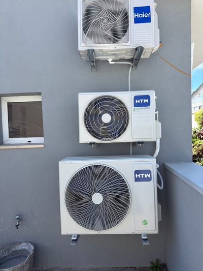 The image shows three outdoor air conditioning units mounted on a grey exterior wall. The topmost unit is a Haier brand, and the two units below it are HTW brand. There is a small, closed window to the left of the units. Below the units, a metal faucet is attached to the wall, and a concrete bucket is placed on the ground. The scene is outdoors, and the wall appears to be part of a building. Based on visual cues in the image, the location is likely in the city of Gondomar, Portugal, where this type of architecture and building materials are common. The image is a static, eye-level shot of the wall and the air conditioning units. There are no people or animals visible. The environment appears to be daytime.