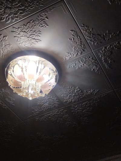 An ornate chandelier with a central light source hangs from a dark, embossed ceiling in Vinogradnoe, Kyrgyzstan. The ceiling panels feature a repeating pattern of stylized floral designs and linear elements, creating a textured, visually complex surface. The chandelier itself has a circular frame containing translucent, petal-like shades and hanging crystal elements. The lighting from the chandelier casts a warm glow upwards and illuminates parts of the embossed ceiling, creating shadows that accentuate the intricate details of the pattern. The overall scene is dimly lit, with the focus on the illuminated chandelier and the patterned ceiling.