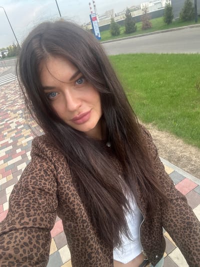 A woman is standing on a paved sidewalk in Edem, Russia. The sidewalk is made of multi-colored rectangular stones. A grassy area with several small trees is adjacent to the sidewalk. A roadway is visible beyond the grassy area. In the background, there is a building with blue and white signage, and industrial structures including smokestacks. The woman is wearing a brown animal print jacket. She has long, dark brown hair and light blue eyes. She is looking at the camera.