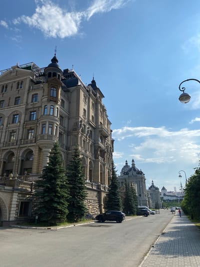 In the city of Bogorodskoe, Russia, on a clear day with scattered clouds, an ornate building with intricate architectural details stands tall on the left side of the frame. The building features multiple stories, bay windows, decorative moldings, and a dark, pointed roof. Several evergreen trees are planted in front of the building. A paved road runs alongside the building, with a sidewalk on the right. Several cars are parked along the road, and a few pedestrians are visible walking on the sidewalk. In the background, more buildings with domed roofs can be seen, along with a distant white tower. A street lamp with a curved arm extends into the sky from the right side of the image.