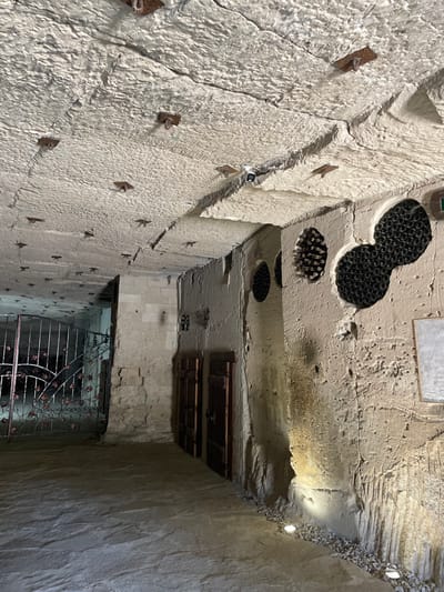 The image depicts an underground passage or cellar within the city of Brănești, Moldova, likely part of a wine storage facility. The environment consists of rough, light-colored, excavated rock walls and ceiling, exhibiting prominent tool marks and natural striations. The ceiling features numerous dark, rusty metal anchor points and a white security camera with a blue indicator light. The floor is paved with large, irregularly shaped, light-colored flagstones.

On the right, a textured rock wall contains multiple carved niches of varying sizes and shapes, including circular and bilobed forms, each densely packed with horizontally stored dark-colored wine bottles. Three ground-level spotlights are embedded in a strip of white pebbles at the base of this wall, illuminating its rough surface. A light-colored rectangular plaque is affixed higher on the wall.

Towards the center-left, a constructed wall section of light-colored, cut stone blocks is visible. This section features two dark brown, wooden doors with visible metal hardware, recessed into the wall. Further back on the left, a large, ornate black metal gate with decorative scrollwork and floral motifs partially blocks a dimly lit passage. No individuals are present, and no actions or interactions are depicted.
