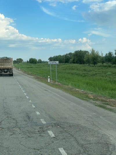 A truck is driving down a cracked asphalt road with white dashed lines. The truck is facing away from the camera. To the right of the road is a sign that says "SUWENLI" and a grassy field with trees in the background. The sky is blue with white clouds. The scene appears to be in a rural area.