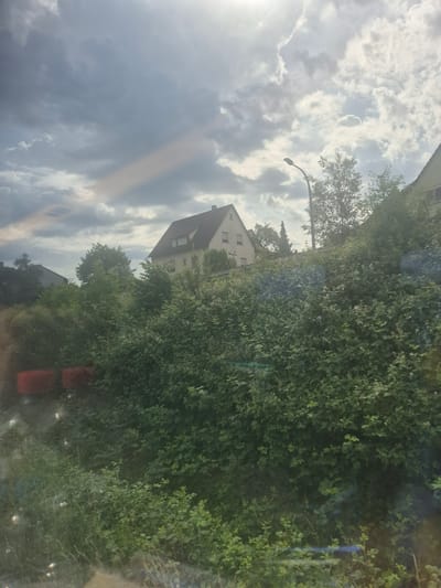 The image shows a view in Neudenau, Germany. On the right is a dense green hedge and trees. In the center, there is a light-colored house with a gray roof. A street light is visible near the house. The sky is filled with gray and white clouds with the sun peeking through, casting a bright ray. The overall composition suggests a suburban or residential area.