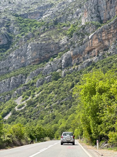 The image shows a silver Toyota Land Cruiser parked on a gray asphalt road alongside a steep, rocky mountainside with sparse green vegetation. The vehicle's license plate reads "PG-HK 482." A white line divides the road. The location is Donje Mrke, Montenegro, as evidenced by the license plate. The road is situated along a drop-off. A road sign is visible near the edge.