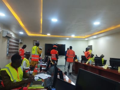The image shows a meeting room in an office building located at latitude 10.929 and longitude 11.476 in Ashaka, Nigeria. There are eight men in the room, all wearing yellow vests and some wearing orange shirts. The men are seated at a long wooden table with laptops, keyboards, monitors, and other office supplies. The room has a white ceiling with recessed lighting, a large flat-screen TV on the wall, and beige walls. Some men are engaged in conversation, while others are working on laptops or talking on their phones. The man in the foreground appears to be writing on a notepad while looking down. The scene suggests a training session or work-related meeting taking place.