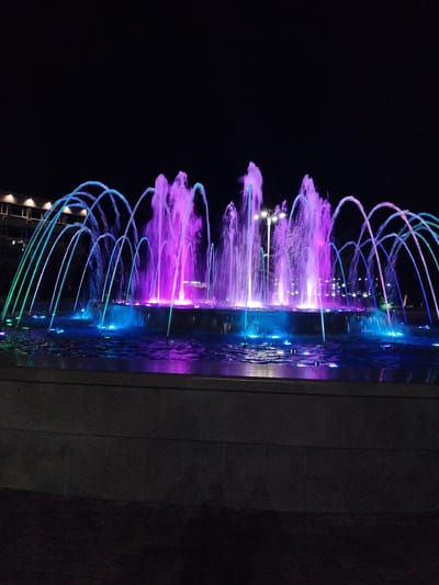 This media file features a vibrant, illuminated fountain at night in Vinogradny, Russia. The fountain is the central subject, with multiple jets of water arcing upwards and outwards. The water is lit with a dazzling array of colors, primarily bright purples and blues, with some green and cyan accents visible on the outer streams. The lights create a mesmerizing spectacle against the dark night sky.
The fountain is situated in a pool of water, which reflects the colorful lights. Submerged lights also contribute to the overall illumination, casting a glow upwards through the water and onto the falling streams. In the background, there's a glimpse of a building with illuminated windows and exterior lights, suggesting an urban or developed setting. The structure of the fountain base is visible, appearing to be made of stone or concrete, with a darker foreground suggesting a paved area or walkway.
The time of day is clearly night, and the sky is completely dark, without any visible stars or moon, indicating a clear or overcast night. There are no people visible in the frame, and the focus is entirely on the dynamic display of the fountain. The overall mood conveyed is one of beauty, tranquility, and perhaps a sense of wonder, due to the artistic play of light and water. No text is visible in the image.