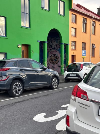 The image shows a street scene in Reykjavik, Iceland, with parked cars in front of brightly colored buildings. The building on the left is painted green with blue-framed windows, and the building above is painted orange with a red roof. There's a mural above a large arched opening on the green building. Parked in front are a dark-colored SUV and two other cars. A blue and white sign for electric vehicle charging is visible attached to the orange building. A white painted arrow is on the street.