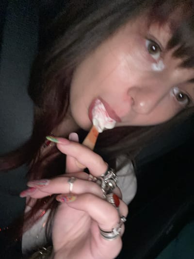 A fair-skinned woman with dark hair and bangs is captured in a close-up shot. Her lips are pursed around a popsicle with what appears to be whipped cream. She is looking off to the side. On her hand, there are multiple rings, some with large stones. Her fingernails are long and painted with a multi-colored design. The background is dark.