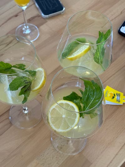 Three glasses of pale yellow drinks with lemon slices and mint leaves sit on a wooden surface. The glasses have stems. One glass contains a small amount of orange liquid, likely from a Mimosa. A yellow tag with the word "Limoncello" is near one of the glasses. A black object with the number "504" on it and part of a smartphone are also visible. The drinks appear refreshing, like lemonade or a similar citrus beverage.
