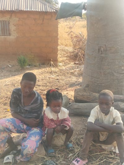 Three children sit outside in a rural setting. An older child wears patterned pants and a patterned top, sitting to the left. A small girl with braided hair sits next to them, wearing a red and white dress. To the right, a child with folded arms wears a light shirt and dark pants. They all appear pensive. Behind them is a mud brick building with a corrugated metal roof and a large tree. The ground is dry and covered with sparse vegetation.