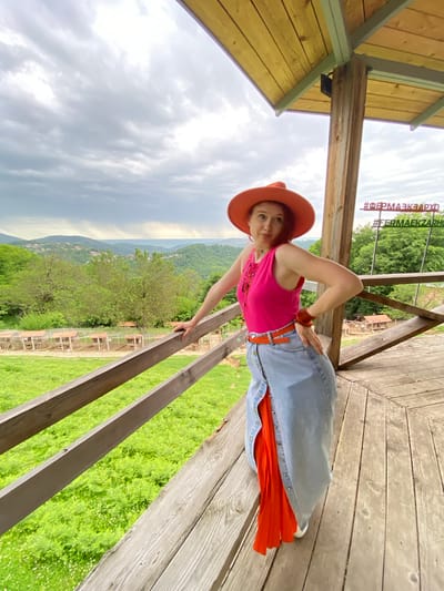 A woman with red hair wearing an orange hat, pink tank top, jeans skirt, and orange belt is posing on a wooden platform with a railing. She is standing on the platform and leaning against the railing with her right hand. She is looking at the camera with a slight smirk. In the background, there is a lush green landscape, including trees, hills, and a cloudy sky. The hashtags #ФЕРМАЭКЗАРХО appear in the upper left. The location is likely in Прогресс, Russia, near a tourist destination or attraction. The woman is possibly engaging in a photoshoot or documenting her visit.
