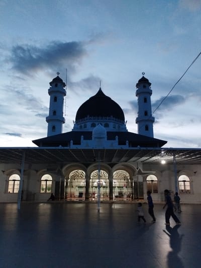 The media file depicts a mosque at dusk, with a dark, cloudy sky overhead. The mosque has a large central dome and two tall minarets, both topped with a crescent moon symbol. The building is illuminated from within, casting a warm glow through arched windows and doorways. In the foreground, a paved courtyard is visible, reflecting the mosque's lights and the twilight sky.
Several people are present in the courtyard. A solitary figure sits on the ground in the shade of an archway. Further into the courtyard, three younger individuals, appearing to be children or teenagers, are walking and playing. Their silhouettes are cast long shadows on the wet-looking pavement.
The scene suggests a peaceful evening atmosphere, possibly after prayer. The location is identified as Mesjid Yaman, Indonesia. There is visible Arabic calligraphy on the arched windows of the mosque, but it is not legible enough to translate. The overall mood is serene and contemplative, with the fading light creating a dramatic backdrop for the majestic mosque.