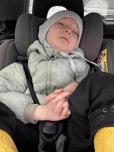 The media file captures a peaceful scene of a baby sleeping soundly in a car seat, likely within a vehicle traveling in Pavlovskii, Russia. The infant is dressed warmly in a light sage green puffy jacket and a light grey ribbed beanie, indicative of cold weather. A blurry, light-colored landscape, possibly snowy, is visible through the car window, reinforcing the winter setting.
The baby is secured in a dark grey car seat with a prominent headrest. An adult's hand, partially visible on the right, gently holds the baby's hands, suggesting comfort and reassurance during the journey. The car seat's safety features are visible, including a black harness strap across the baby's lap. A yellow warning label on the car seat headrest clearly shows the text "AIRBAG" and symbols advising against placing a child seat in front of an active airbag. A small, colorful charm, possibly a cartoon character, is attached to the zipper of the baby's jacket. The baby's closed eyes and relaxed posture convey a sense of deep sleep and contentment.
