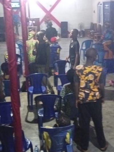 People are gathered indoors, sitting on and standing near blue plastic chairs. Some chairs are empty. A speaker and a fan are in the background. Red and white decorations are visible on the wall. The lighting is dim. The scene seems to depict a gathering of some sort.
