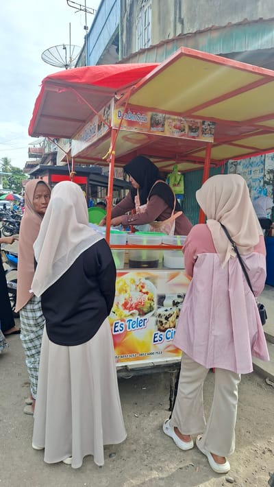 In Keudegeudong, Indonesia, a street-side food stall is busy serving customers. The stall is adorned with a red and yellow awning and displays colorful advertisements for various Indonesian desserts, including "Es Teler," "Es Campur," and "Es Sirup Cincau." The text on the stall, "Doa Dek Syila," likely indicates the name of the business.
Several individuals are visible, primarily women wearing hijabs. One woman, in a pink striped top and light-colored pants, is standing near the stall. Another woman, wearing a black hijab and a pink top, is actively preparing a drink behind the counter, reaching into containers filled with ingredients. A third person, in a beige hijab and a patterned skirt, is seated closer to the foreground. The scene appears to be set during the daytime, under what seems to be bright or sunny weather, given the lighting. The background suggests an outdoor, urban environment with buildings and parked motorcycles. The overall impression is of a lively, everyday commercial activity at a local food vendor.
