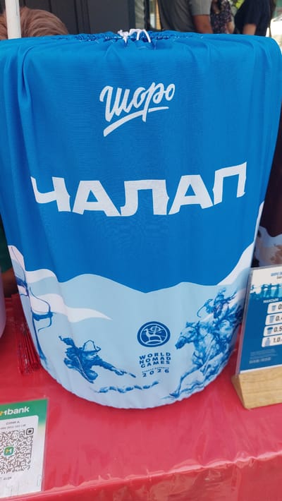 This media file shows a close-up of a blue fabric object, possibly a bag or a cover, with white lettering. The object is draped over a red surface, likely a table. On the left side of the fabric, there's a white word written in a stylized script. Prominently displayed on the fabric is a large, white word, "ЧАЛАП," written in a bold, sans-serif font, which appears to be a place name or a brand.

Further to the right, there is a circular emblem with a depiction of a person engaged in some activity, surrounded by text that reads "WORLD NOMAD GAMES 2026" and "KYRGYZ REPUBLIC". The fabric also features a blue and white graphic design with swirling patterns that resemble water or clouds, and some stylized figures.

In the background, out of focus, are people, suggesting a public event or gathering. A corner of a wooden stand with some printed information, including what looks like a price list with numbers and abbreviations like "MAXIM" and "SONYA A.", is also visible. At the bottom right, a green card or label with a QR code and the text "Mbank" is partially visible.

The overall impression is that of a promotional item or merchandise related to the "World Nomad Games 2026" being held in the Kyrgyz Republic, possibly in or near a location associated with "Чалап". The setting appears to be outdoors or in a casual, perhaps market-like environment, given the red table and other people in the background.