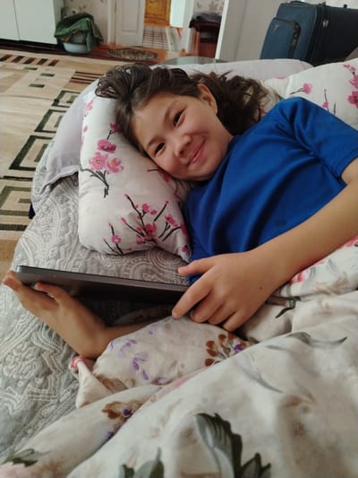 A young girl with dark hair is lying in bed, holding a tablet computer. She is smiling and looking at the camera. The room is decorated with floral patterns on the bedding and pillows. In the background, there is a rug on the floor and two suitcases. This appears to be an indoor setting, possibly a bedroom in a home in Barskoon, Kyrgyzstan.