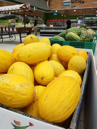 This media file captures a close-up view of a display of ripe yellow melons in a supermarket produce section. Several large, oblong melons are piled high, their smooth, bright yellow skin showing subtle variations in tone and some minor blemishes. In the foreground, the rounded shapes of the melons dominate the frame, with some slightly deflated ones at the bottom of the display. The melons are arranged in a crate with weathered blue and natural wood accents, suggesting they are part of a larger bulk display.

Behind the melons, a glimpse of the supermarket environment is visible. In the mid-ground, a green plastic crate holds a collection of watermelons, their dark green rinds contrasting with the yellow of the foreground fruit. Further back, the store's interior is illuminated by overhead lighting, creating a bright, commercial atmosphere.

In the background, several people are present, casually browsing the aisles. One person, wearing a black t-shirt and dark pants, is visible at a distance, looking towards the produce. Two women, one in a white top and the other in a black top and green pants, are further back, near a wooden display stand. This wooden stand has a white canopy and appears to be selling pre-packaged goods, with visible price tags of 2, 5, 9, and 18.

Above the produce section, signage is visible. Text in Arabic, including "الكيلو" (Al-Kilo - The Kilo) and "الجزار" (Al-Jazaar - The Butcher), is displayed on wooden boards, indicating different sections of the supermarket. The overall impression is a typical grocery shopping scene, likely during daytime hours given the bright interior lighting. The focus remains on the abundance and freshness of the fruit.