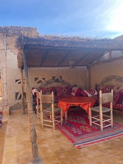 An outdoor seating area features a thatched roof structure against a clear blue sky. Ornate metalwork adorns the walls behind cushioned seating. A small table covered with an orange cloth sits between two chairs on a vibrant red rug. The flooring is tiled in a warm tone, and natural elements like bamboo and dried reeds are used in the building's construction.