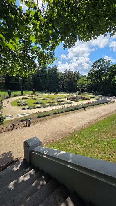 The image depicts a view of a manicured garden with formal flower beds arranged in geometric patterns. The garden is situated in a park with mature trees bordering the flower beds and in the background. Several pathways wind through the garden, and there are white benches along one of the paths. In the foreground, stone stairs and a railing lead down from a vantage point, with dappled sunlight filtering through the leaves of an overhead tree. The sky above is a bright blue with scattered white clouds. A few individuals are visible in the garden, some walking and others standing. This scene is located in the botanical garden of Palanga, Lithuania, specifically the central part of the park with its characteristic landscaping.