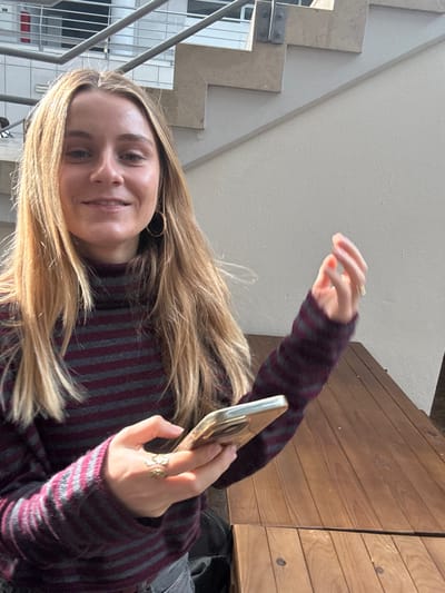 A young adult female is depicted in a well-lit indoor or semi-indoor environment in Porto Salvo, Portugal. She possesses long, straight, blonde hair and fair skin, and is smiling directly at the camera. She wears a horizontally striped dark grey and maroon turtleneck sweater, large gold-colored hoop earrings, and multiple rings on her left hand, including an intricate design on her ring finger. Her left hand holds a smartphone with a light-colored case. Her right hand is slightly raised, with fingers loosely curled and fingernails painted orange. She is positioned at a dark brown, slatted wooden table. In the upper background, a light-colored stone or concrete staircase with a metal railing is visible, adjacent to an off-white wall.