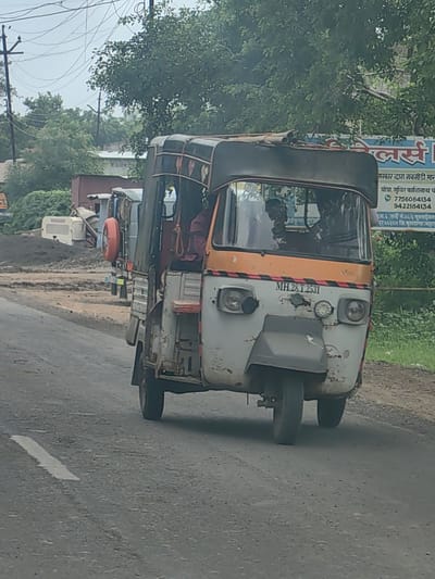 An auto-rickshaw, license plate MH.28.T.2531, is driving on a paved road in Malkapur, India. The auto-rickshaw has a weathered, off-white and orange exterior. It is a three-wheeled vehicle with a covered passenger compartment and a driver's seat. The auto-rickshaw appears to be carrying passengers, though they are not clearly visible. The background shows a construction site with dirt piles and some buildings, as well as lush green trees and overhead power lines. The sky is overcast. The road has a white painted line marking the lane. The scene suggests a typical day in a developing area of Malkapur, with ongoing construction and local transportation.