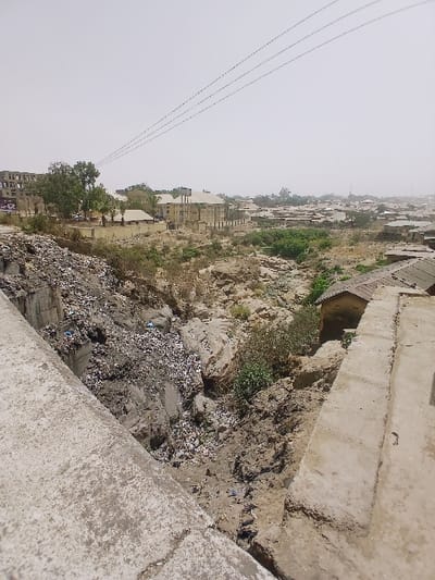 The image displays an elevated view of an urban environment in Kadoma, Zimbabwe. In the immediate foreground, a weathered concrete structure, consistent with a bridge railing, extends diagonally from the lower left. Below this structure, a significant gully or ravine is visible, its left slope heavily filled with accumulated solid waste, including plastics, paper, and various debris. The gully's lower sections consist of dry, irregular rock formations and earthen surfaces, interspersed with sparse vegetation.

On the right side, another concrete structure, possibly a wall or pathway, descends towards the gully. A small, single-story building with a light-colored exterior and a brown, low-pitched roof is partially visible near the gully's edge on the far right.

The midground and background depict an extensive urban landscape under a hazy, light-gray sky. Numerous buildings of varying sizes and architectural styles are present, predominantly multi-story structures with light-colored facades. Dry, sparse trees and bushes are scattered throughout the built environment and within the gully itself. Three parallel utility lines traverse the upper portion of the sky. No individuals or specific activities are observable within the scene. The environment appears dry and extensively developed.
