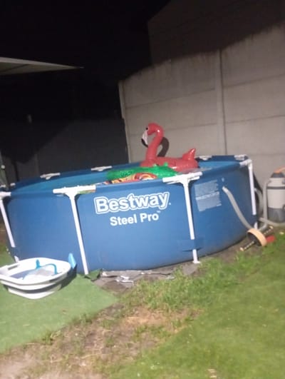 In Kobyłka, Poland, an above-ground swimming pool, branded "Bestway Steel Pro," is positioned on a patch of grass. The pool, filled with water, contains a red inflatable flamingo and a green inflatable alligator. To the left of the pool is a white and blue foldable pool accessory containing a blue net. In the background, a beige concrete fence separates the property from the surrounding area. To the right of the pool, a white and grey water filter is visible, connected to the pool by a hose. The scene appears to be illuminated by artificial light, suggesting it is nighttime or dusk.
