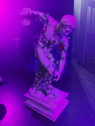 The image shows a statue of a discus thrower, possibly a replica of "The Discobolus," adorned with artificial ivy and red roses. The statue is illuminated by vibrant purple stage lighting, creating a dramatic and moody atmosphere. The statue stands on a multi-tiered pedestal on a dark, reflective floor. In the background, a dark, slightly blurred doorway or partition is visible, suggesting an indoor setting. A sign with the word "Telephone" is faintly visible on the left side of the image. The overall scene suggests a theatrical or artistic display, potentially within a venue like the Wacken Open Air in Kirrwiller, France, though this is speculative.