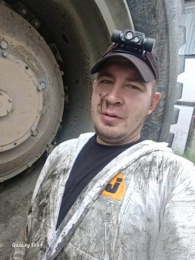 A man is shown in a close-up, possibly a selfie, in front of a large, industrial tire. He is wearing a dirty white protective suit over a black t-shirt, a baseball cap with a headlamp, and has a smudge of dirt on his face. The setting appears to be an outdoor industrial or construction site, with a portion of a building visible in the background. The man's clothing and the surrounding environment suggest he has been engaged in manual labor. The presence of the large tire indicates a connection to heavy machinery or vehicles. The man's gaze is directed towards the camera, and his expression is neutral. The overall scene conveys a sense of hard work and an industrial setting.