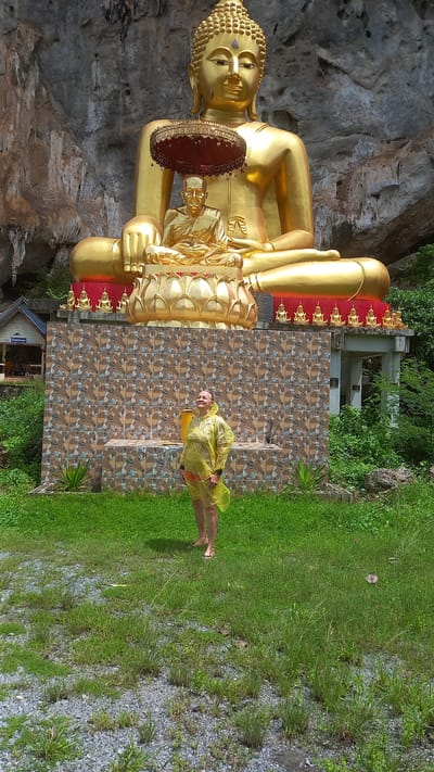 A wide-angle shot of a golden Buddha statue seated within a cave at Wat Tham Ta Pan Temple in Thap Put, Phang-nga, Thailand. A person in a yellow raincoat stands barefoot to the right of the statue in a grassy area. A mosaic tile wall and patches of green vegetation are present near the statue. The ground is covered with green grass and grey gravel.