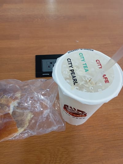An overhead shot captures a snack on a wooden surface, likely a table or desk, in what appears to be an indoor setting. No people are visible.

The primary subjects are a large white disposable cup with a straw and a pastry or bread in a clear plastic bag. The cup, showing condensation on its exterior, suggests a cold beverage. The cup's lid prominently displays "CITY TEA" and "CITY PEARL" in green and black text, along with "CITY CAFE" in red, indicating the brand. Chinese characters on the lid, "撕開即飲就口喝少用吸管愛地球," translate to an eco-friendly message: "Tear open and drink directly, use fewer straws to love the Earth." The liquid inside the cup is light-colored and milky, consistent with a pearl milk tea or similar beverage. The lower part of the cup features a red "CITY CAFE" logo with Chinese text, "在城市 探索城" (In the city, explore the city).

Next to the cup, a piece of bread or pastry is enclosed in a clear plastic bag. It appears golden-brown with sliced almonds on top and seems partially eaten or broken.

In the upper left background, a black Panasonic power outlet is embedded in the wooden surface, featuring two Type A/B sockets. This detail, combined with the visible Chinese text, reinforces the location context of Kaohsiung, Taiwan, where such outlets are common. The scene suggests a casual break, perhaps at an office, cafe, or study area.