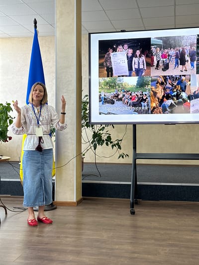 A woman stands in front of a large screen displaying a collage of photos. The screen shows various images of people engaged in different activities. The woman is wearing a traditional embroidered shirt, jeans, and red shoes. She is gesturing with her hands as if speaking or presenting. Behind her, a Ukrainian flag is visible. The scene appears to be indoors, possibly a conference room or auditorium, with wooden flooring and beige walls. Plants are present in the foreground. The overall mood seems to be one of presentation or education. The visible text on the screen reads "Процес залучення". The location is Myronivka, Ukraine.