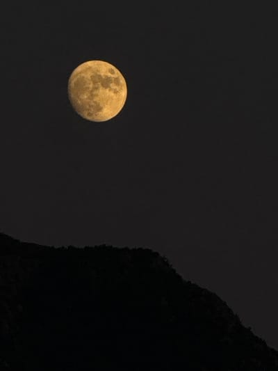 The image depicts a full moon in a dark, starless sky. The moon is illuminated with a yellowish hue and shows visible surface features. In the foreground, a dark, silhouetted mountain ridge runs across the bottom of the frame. The scene is devoid of any signs of human presence, indicating a natural landscape at night. The location is identified as the city of Trap, Montenegro.