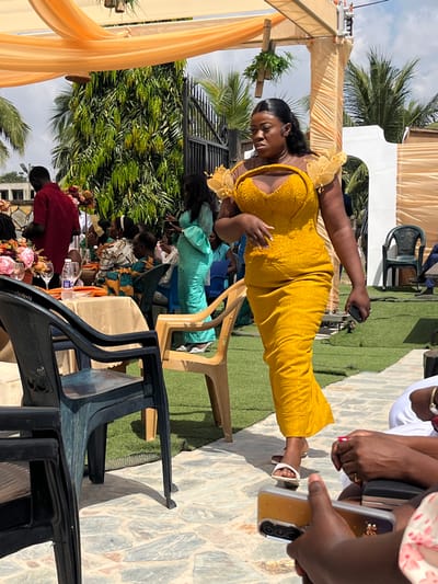 The image captures an outdoor social event under bright daylight in Kumasi, Ghana. The scene features multiple individuals gathered beneath light orange fabric canopies adorned with green foliage.
In the mid-ground, a woman with dark skin, styled dark hair, wears a vibrant mustard yellow traditional-style dress. The dress features a beaded off-shoulder sweetheart neckline bodice with ruffled sheer yellow fabric detailing on the shoulders. She is walking from left to right across a ground composed of irregular flagstones and artificial green turf, holding a dark-colored smartphone in her left hand. She wears white flat sandals and appears to be looking towards the right side of the frame.
To the left, several people are visible. A man in a dark red long-sleeved shirt stands near a round table covered with a beige tablecloth. The table displays a prominent pink floral centerpiece, empty glasses, and a plastic water bottle. Multiple individuals, some in traditional Ghanaian fabrics, are seated at this table and in the background. A woman in a light blue traditional dress is seen standing further back, holding a smartphone.
The background reveals a large, dark metal gate, dense green foliage, and palm trees under a clear sky. Various plastic chairs in dark blue/green, beige, and black are distributed throughout the event space, some occupied and some empty. In the immediate foreground, the hands of two individuals are partially visible, holding smartphones, one of which has a clear case and a triple camera lens array.