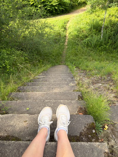 The image shows a first-person perspective of someone standing on a concrete staircase in a natural outdoor environment. The stairs are worn and partially overgrown with grass and moss. The viewpoint is looking down the stairs, which recede into the distance and are flanked by dense green foliage. A dirt path is visible at the top of the stairs, winding through grassy areas. The person is wearing white athletic shoes with white socks and visible legs. The lighting suggests it is daytime with sunlight filtering through the trees. This scene is located in the vicinity of Tēraudi, Latvia.