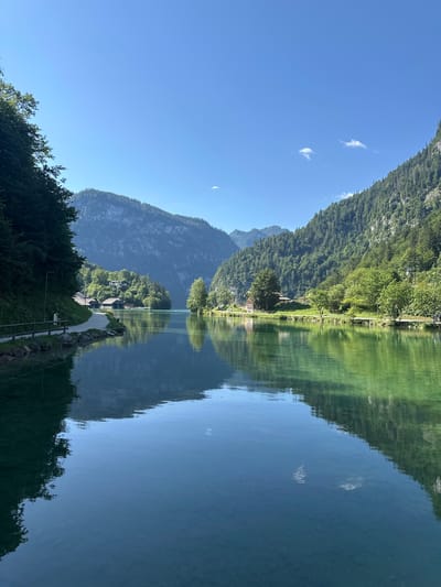 The image depicts a tranquil scene at the Königssee lake in Schönau am Königssee, Germany. A clear blue sky stretches above lush green mountains that flank the serene, reflective body of water. The shoreline features a paved walkway lined with trees and a wooden fence, with a few rustic buildings nestled amongst the greenery. The water's surface acts as a mirror, reflecting the surrounding landscape with striking clarity. The overall impression is one of peaceful natural beauty, with the architecture seamlessly integrated into the environment.