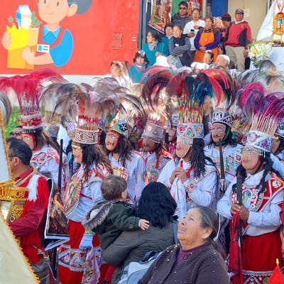In Wanchaq, Peru, a cultural event unfolds with participants dressed in traditional festive attire. Several individuals are adorned in elaborate costumes featuring tall, feathered headdresses and masks with painted faces. These figures, likely part of a procession or performance, are positioned in the foreground and midground. Behind them, a diverse crowd of spectators watches the proceedings, with varying expressions and stances. The backdrop reveals a red wall with decorative elements and hints of buildings and banners, suggesting an urban setting within the city. A young child is visible, being held by an adult, and an elderly woman in the foreground looks upwards, her expression one of engagement with the event. The scene captures a moment of cultural celebration, with a strong emphasis on the colorful and ornate traditional costumes.